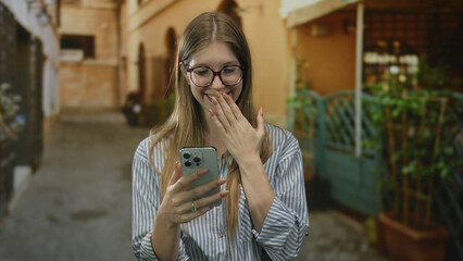 Obraz premium Woman covers mouth with hand while looking at smartphone screen on a sunlit old town street; surprise.