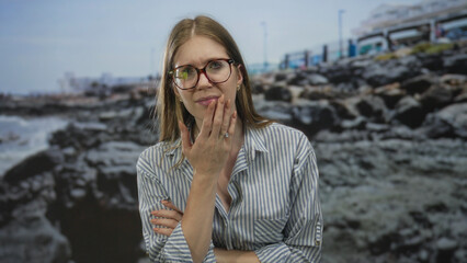 Blonde woman wearing glasses and striped shirt with hand on cheek by rocky seaside beach; doubt...