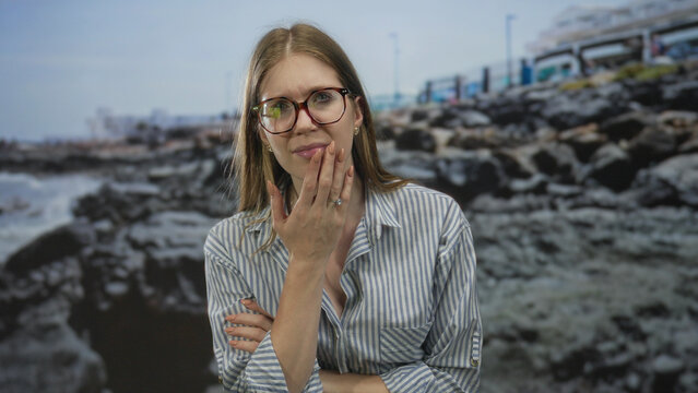 Blonde woman wearing glasses and striped shirt with hand on cheek by rocky seaside beach; doubt concern thought.