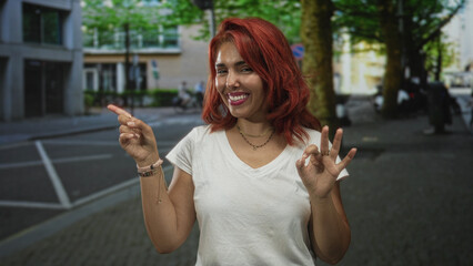 Young hispanic woman points finger and makes ok sign on city street while smiling with closed eyes; joy confidence.
