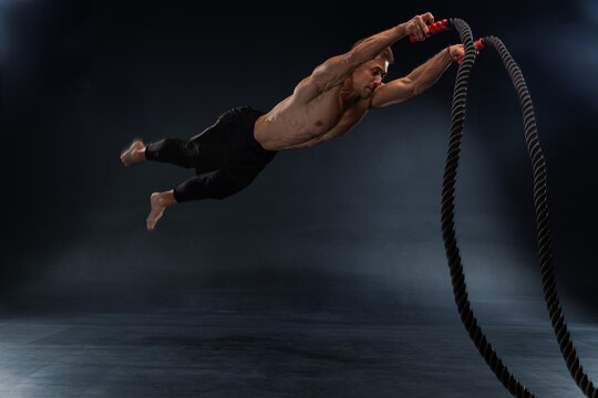 muscular man is leaping off the ground while gripping heavy battle ropes. He is in mid air showcasing strength and determination in a dark training studio setting. - Powered by Adobe