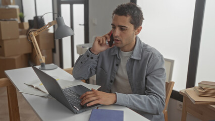 Young hispanic man talking on phone while typing on laptop in modern home office with desk lamp and books around.
