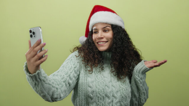 Woman wearing santa hat makes video call over yellow background, dressed in cozy sweater, exuding holiday cheer and joy through smartphone, isolated indoors. - Powered by Adobe