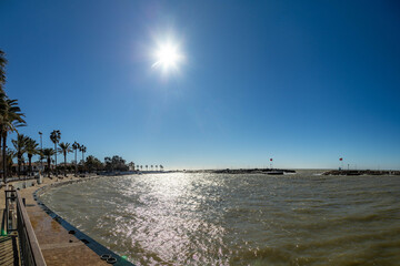 Empty marina glistens in stormy sunlight under a clear blue sky. Traditional houses with tile roofs line the palm-filled promenade as flags wave over the dark sea. Side, Antalya, Turkey, Mediterranean