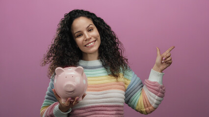 Woman happily points sideways holding a pink piggy bank against an isolated pink background, wearing a colorful striped sweater, symbolizing savings and financial planning.