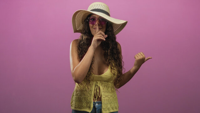 Hispanic woman in straw sunhat and purple sunglasses points thumb to side then makes finger to lips shush in studio; playful secrecy. - Powered by Adobe