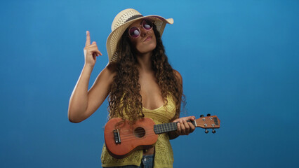Woman strumming ukulele in studio wearing wide sun hat and purple sunglasses, holding and motioning with raised right hand while smiling; playful summer fun.