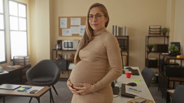 Pregnant blonde woman smiling in an office setting with desks and computers, showcasing a modern workplace environment