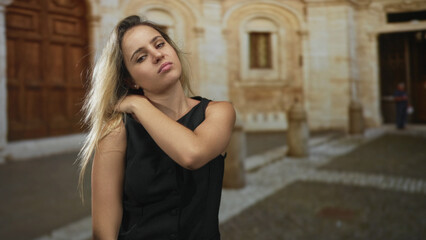 Fototapeta premium Woman with hand on neck revealing bare shoulder in old town building courtyard with stone archways; melancholy.