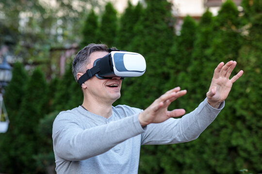 Happy middle-aged man wearing a virtual reality headset, reaching and gesturing in a virtual world against a background of green bushes