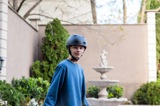 Young teenage boy in a black safety helmet and blue sweater standing outside, looking at the camera with a serious expression near a garden fountain