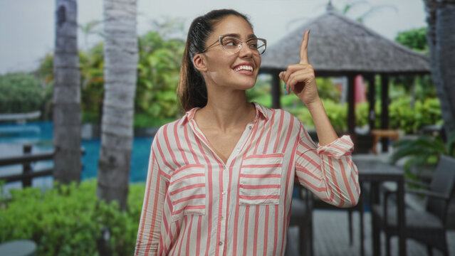 Woman wearing glasses and striped shirt pointing finger at building near pool at resort by palm trees; inspiration optimism.