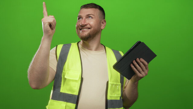 Man engineer in high visibility vest pointing finger while holding tablet in green studio; confidence planning.