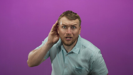 Man cupping hand to ear and leaning forward in purple studio wearing light blue shirt; curiosity attention.