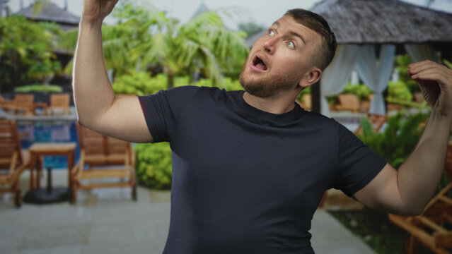 Man with hands raised and open mouth gesture at resort pool beside hotel building and cabana lounge chairs; surprise vacation leisure.