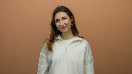 Obraz premium Young woman smiling confidently against isolated brown background wearing casual white sweater showcasing relaxed demeanor.