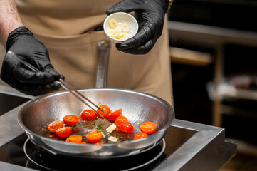 Professional chef in black gloves cooking cherry tomatoes and sliced garlic in a stainless steel pan on a stove for an Italian dish like pasta sauce.