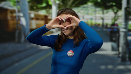 Fototapeta premium Woman wearing vote badge forms heart shape with hands on city street smiling in sunlight during us election outdoors showing patriotic support