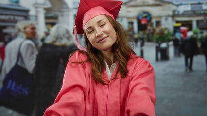 Woman in pink cap and gown celebrating graduation on city street with joyful expression surrounded by urban life and people passing by in background outdoors