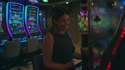 Young hispanic brunette woman shouting and cheering with clenched fists at a slot machine row inside a building with neon lights; winning joy.