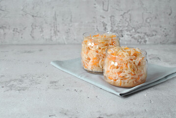 Two small glass jars filled with shredded fermented cabbage and carrots placed on a folded light gray napkin. Minimalist, clean kitchen setting with soft natural lighting and a neutral concrete backgr