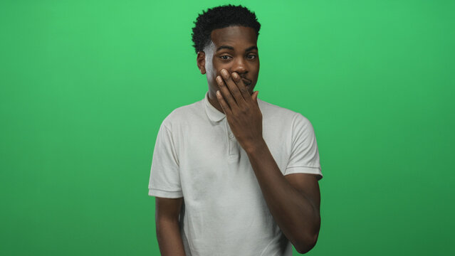 Young man in white shirt posed covers mouth with hand against vibrant green studio backdrop; surprise.