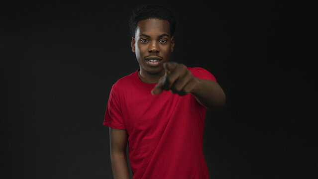 Friendly smiling young african american man pointing finger in studio against black wall wearing red shirt; confidence.