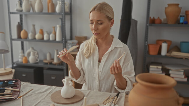 Woman wearing white blouse holding carving tool at pottery table with sculpted vase in studio; serenity.