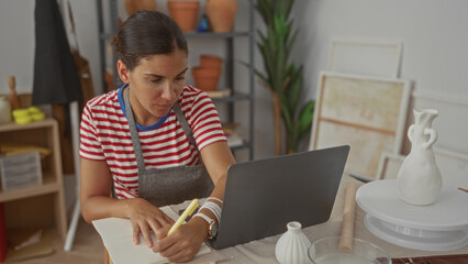 Woman sketching with pencil, hand on sketchbook at pottery studio table with laptop and vases;...