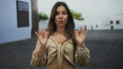Fototapeta premium Woman makes ok sign with hands and exposed midriff on a street with white buildings; calm meditation.