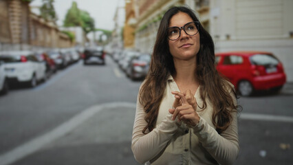 Woman with glasses on a city street, hands clasped with fingers interlaced while looking upward near parked cars and buildings; thoughtful.