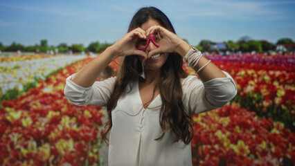 Obraz premium Woman making heart with hands and wearing pink glasses while smiling and standing among red tulips in front of building and houses under blue sky; love joy.