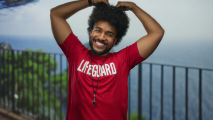Lifeguard man smiling by the seaside promenade with heart gesture, wearing red uniform, against the...