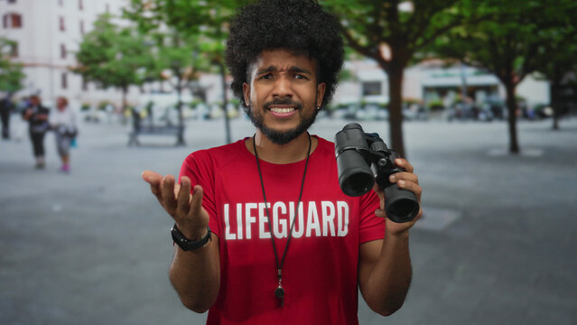 Lifeguard man with afro hairstyle holding binoculars, expressing frustration on a city street with trees and buildings in the background.