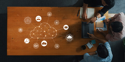 A top-down view of a wooden table where people collaborate using laptops and tablets, surrounded by cloud computing icons symbolizing digital connectivity and teamwork. Trope