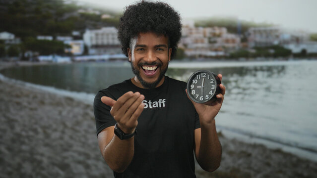Smiling man with afro hair in staff uniform holding a clock, extends his hand towards the camera with a seaside backdrop of sand, water, and buildings in view. - Powered by Adobe