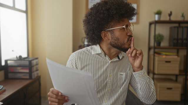 Man reading papers in office setting with focused expression, wearing glasses and striped shirt, while gesturing for silence, surrounded by organized shelves and plants in the background.