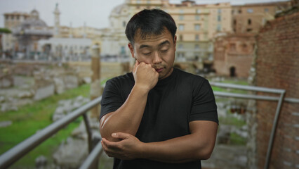 Fototapeta premium Young chinese man with hand on cheek resting elbow at weathered building facade among ancient stone ruins; contemplation.