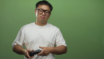 Young chinese man wearing glasses holds gamecontroller and moves left hand in green studio;...