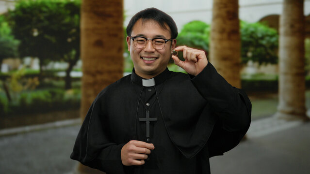 Asian priest holding a ring with a joyful smile stands outdoors in a church courtyard, symbolizing marriage and religious commitment.