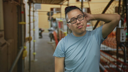 Young chinese man with hand on chin in building scaffolding aisle at construction site outdoors; thoughtful reflection.