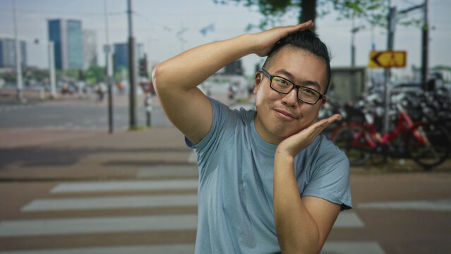 Man with glasses in blue t shirt smiling with hands framing face on street crosswalk beside parked bicycles and city buildings; playful.