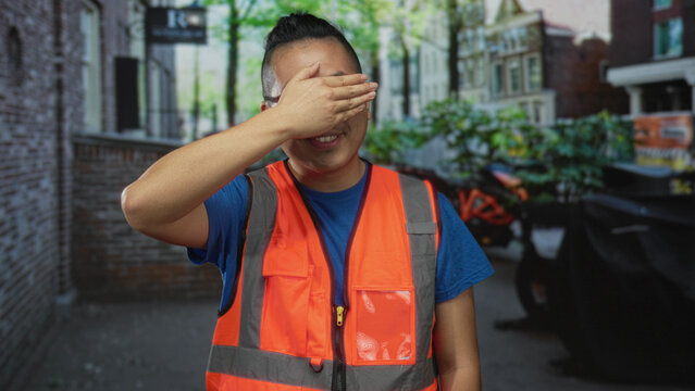 Young man wearing orange reflective vest raising hand to forehead shielding eyes on a city street beside parked motorcycles; fatigue duty.