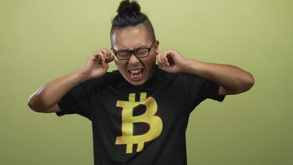 Man wearing bitcoin t shirt plugs fingers in ears in studio with olive green backdrop; annoyance silence.