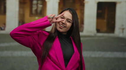 Fototapeta premium Woman smiling outside historic university wearing pink suit and glasses with a peace sign gesture, capturing youthful energy in an educational setting.