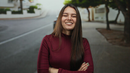 Fototapeta premium Woman smiling with arms crossed standing on an outdoor street, showcasing a confident and relaxed expression in a casual urban environment.