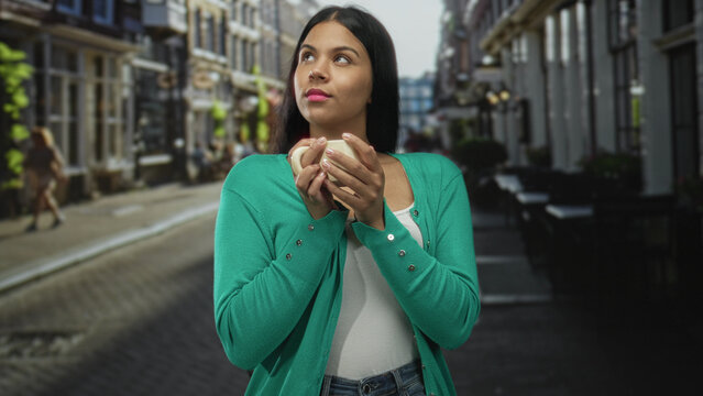 Young latin woman wearing green cardigan holding coffee cup on city street looks up with thoughtful expression; quiet contemplation.