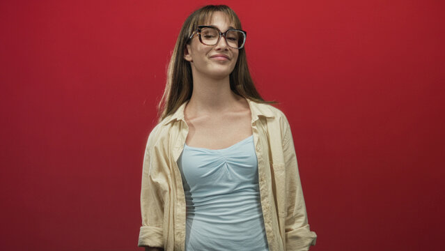 Woman points finger upward in red studio with glasses and cream buttondown over light blue top, visible hand gesture and rolled sleeve; confidence attitude.