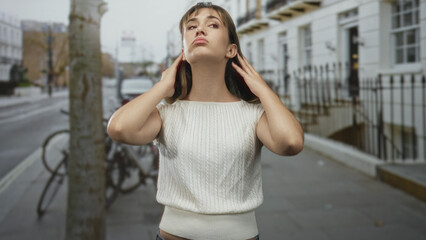 Woman touching hair with hands on city street wearing white sweater and jeans, gaze upward and fingers near face; serenity introspection.