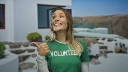 Young blonde woman outdoors giving a thumbs up, wearing a volunteer shirt, expressing positivity in an urban street setting with a blurred cityscape in the background.